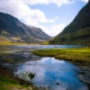 Breathtaking view of a valley with a reflective lake in the Scottish Highlands under a sunny sky.