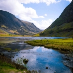 Breathtaking view of a valley with a reflective lake in the Scottish Highlands under a sunny sky.